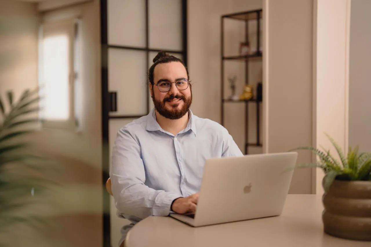 Marc J. Cabrer en retrato editorial, con camisa azul claro, gafas y barba, en un entorno cálido.