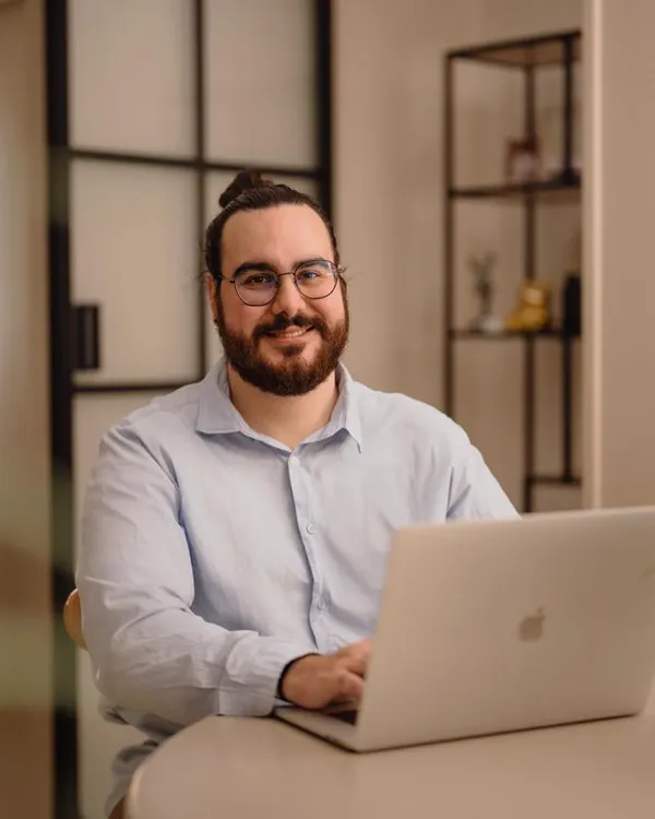 Retrato de Marc J. Cabrer sentado en una mesa con su ordenador portátil. Lleva gafas, barba y una camisa azul claro, en un ambiente cálido e iluminado con plantas decorativas.